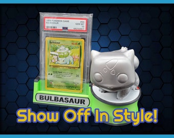 May include: A green and black display stand with a silver Bulbasaur figurine and a graded Pokemon card in a protective case. The card is a 1999 Bulbasaur card graded Gem Mint 10. The stand has the text "Bulbasaur" and "Show Off In Style!" printed on it.