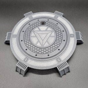 May include: A gray circular stand with a raised, textured center featuring a white Iron Man symbol. The stand has six raised, rectangular sections around the edge.