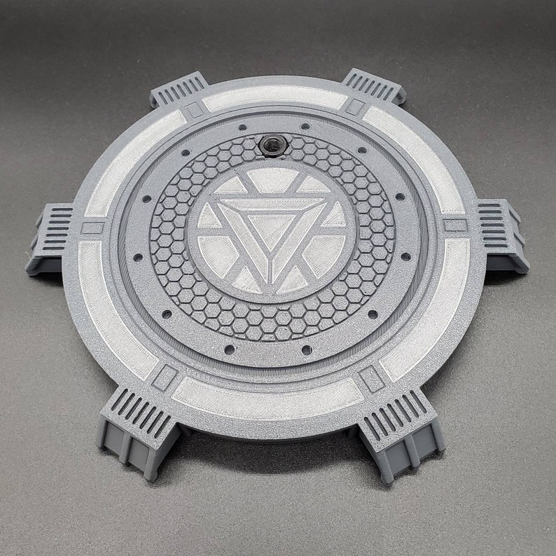 May include: A gray circular stand with a raised, textured center featuring a white Iron Man symbol. The stand has six raised, rectangular sections around the edge.