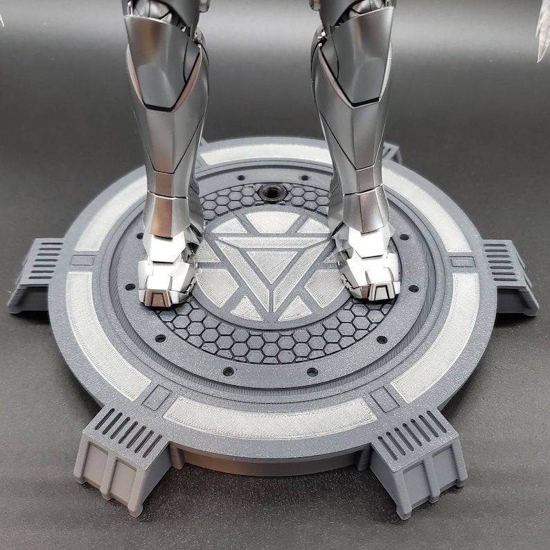 May include: A gray circular display stand with a raised center featuring a stylized Iron Man symbol. The stand has a textured surface and four raised supports.