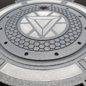 May include: A silver and gray 3D printed round disc with a raised, detailed Iron Man logo in the center. The disc has a textured, hexagonal pattern around the logo.