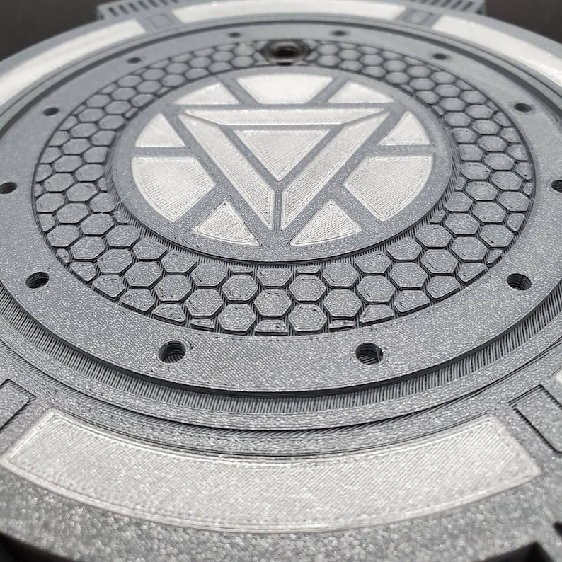 May include: A silver and gray 3D printed round disc with a raised, detailed Iron Man logo in the center. The disc has a textured, hexagonal pattern around the logo.