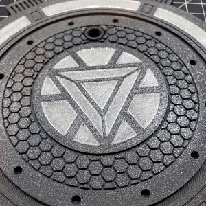 May include: A black and silver round metal plate with a raised hexagonal pattern and a silver Iron Man symbol in the center.