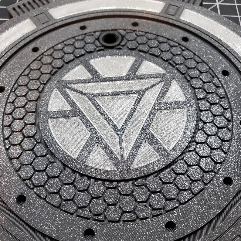 May include: A black and silver round metal plate with a raised hexagonal pattern and a silver Iron Man symbol in the center.