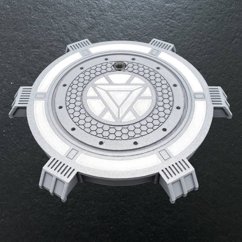 May include: A gray, circular, 3D printed object with a white Iron Man symbol in the center. The object has a textured surface and a raised rim around the edge. It has six legs that extend from the bottom of the object.