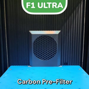 May include: A black square-shaped air filter with a honeycomb-shaped grill. The filter is labeled "Carbon Pre-Filter" and is mounted on a blue surface. The text "F1 ULTRA" is visible in a green circle above the filter.