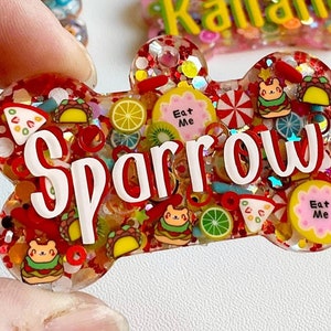 May include: A bone-shaped pet tag with the name "Sparrow" in white lettering. The tag is filled with red glitter and small food-themed charms, including tacos, pizza slices, and "Eat Me" labels.