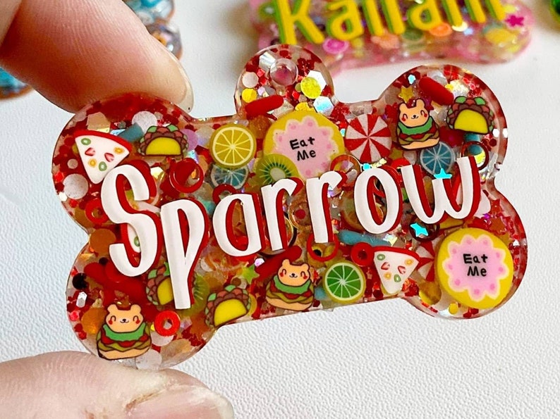 May include: A bone-shaped pet tag with the name "Sparrow" in white lettering. The tag is filled with red glitter and small food-themed charms, including tacos, pizza slices, and "Eat Me" labels.