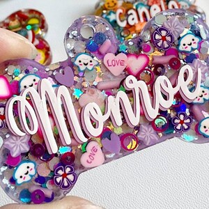 May include: A purple, bone-shaped resin charm with the name "Monroe" in white script. The charm is filled with glitter, hearts, flowers, and smiling cloud and star shapes. The word "LOVE" is on a pink heart.