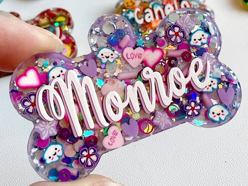 May include: A purple, bone-shaped resin charm with the name "Monroe" in white script. The charm is filled with glitter, hearts, flowers, and smiling cloud and star shapes. The word "LOVE" is on a pink heart.