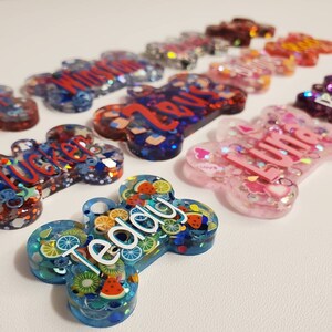 May include: A collection of colorful, bone-shaped pet tags. The tags are made of resin and feature names like "Teddy," "Tucker," and "Luna." Some tags have fruit slices, stars, and glitter. The tags are in shades of blue, pink, red, and purple.