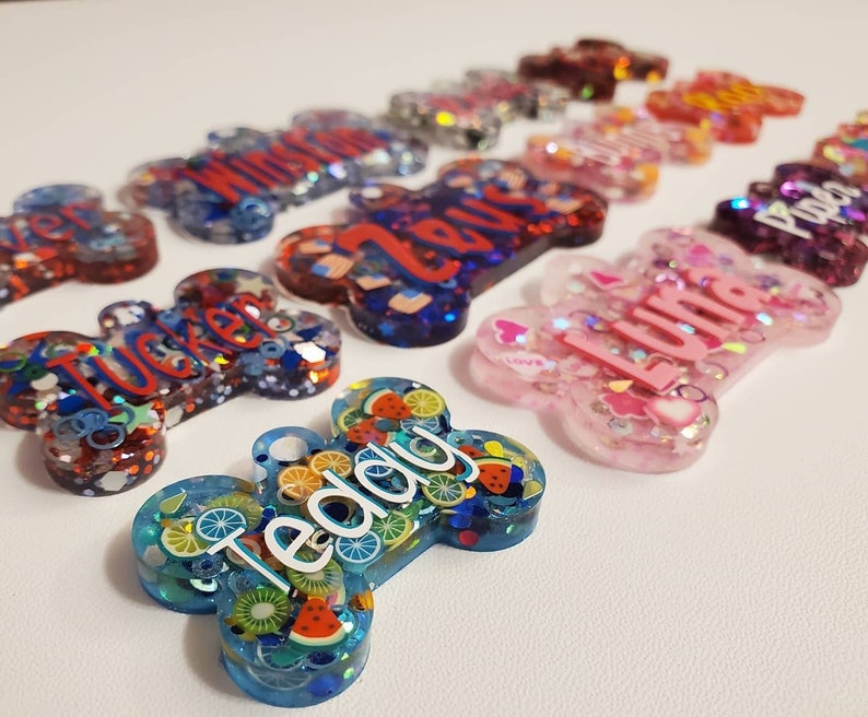 May include: A collection of colorful, bone-shaped pet tags. The tags are made of resin and feature names like "Teddy," "Tucker," and "Luna." Some tags have fruit slices, stars, and glitter. The tags are in shades of blue, pink, red, and purple.
