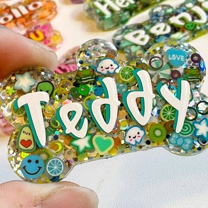 May include: A bone-shaped pet tag with the name "Teddy" in white and teal lettering. The tag is filled with glitter and colorful charms, including smiley faces, fruit slices, and hearts. Other pet tags are visible in the background.