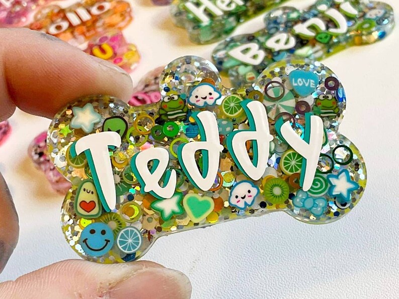 May include: A bone-shaped pet tag with the name "Teddy" in white and teal lettering. The tag is filled with glitter and colorful charms, including smiley faces, fruit slices, and hearts. Other pet tags are visible in the background.