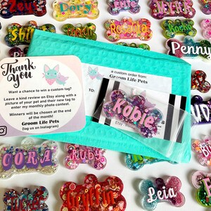 May include: A collection of colorful, bone-shaped pet tags with names like "Kobie," "Penny," and "Leia." The tags are surrounded by glitter and decorative elements. A teal bubble mailer and a thank you card from Groom Life Pets are also visible.