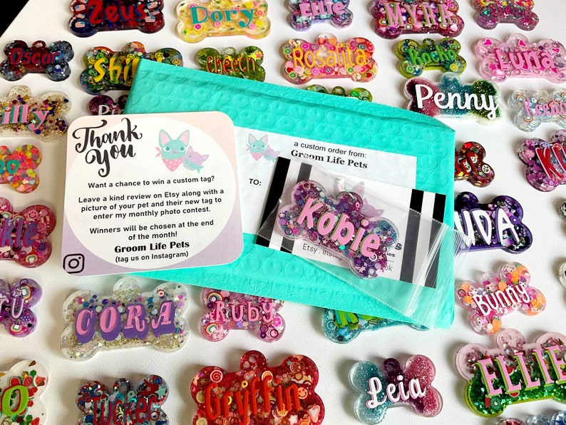 May include: A collection of colorful, bone-shaped pet tags with names like "Kobie," "Penny," and "Leia." The tags are surrounded by glitter and decorative elements. A teal bubble mailer and a thank you card from Groom Life Pets are also visible.