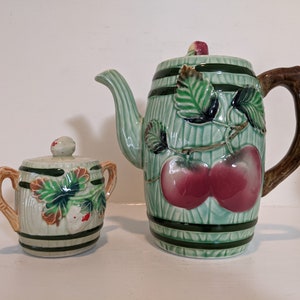 May include: A green and white ceramic teapot and sugar bowl set with a fruit design. The teapot has a brown wooden handle and the sugar bowl has a lid.