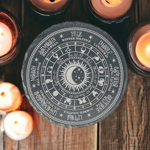 May include: A black slate circular coaster with a white engraved design of a sun and moon with zodiac symbols and dates for the eight sabbats of the Wheel of the Year. The text includes: Yule, Winter Solstice, Imbolc, Candlemas, Ostara, Beltane, Spring Equinox, Litha, Midsummer, Lughnasadh, Mabon, Fall Equinox, Samhain, Halloween.