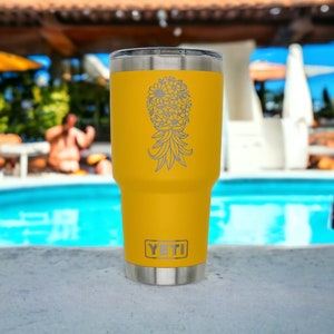 May include: A yellow insulated tumbler with a silver rim and base. A pineapple design made of flowers is etched on the front. The brand name "YETI" is displayed at the bottom. The tumbler is sitting on a concrete surface near a pool.