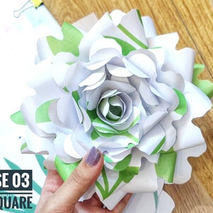 Op de afbeelding: Een witte papieren roos met groene bladeren, de roos is gemaakt van meerdere lagen papier en heeft een gekrulde rand. De tekst "ROSE 03 LA SQUARE" is zichtbaar in de linkerbenedenhoek van de afbeelding.