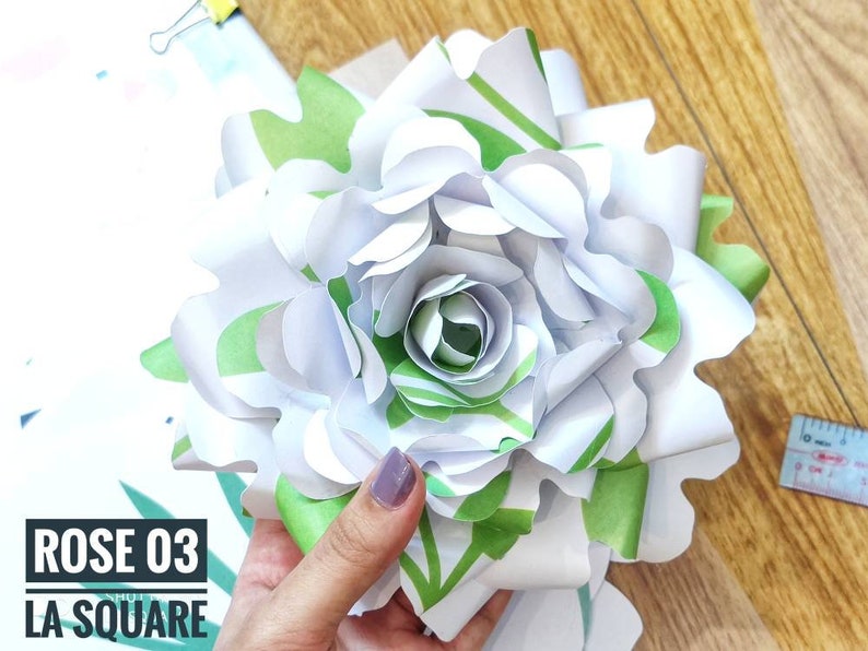 Op de afbeelding: Een witte papieren roos met groene bladeren, de roos is gemaakt van meerdere lagen papier en heeft een gekrulde rand. De tekst "ROSE 03 LA SQUARE" is zichtbaar in de linkerbenedenhoek van de afbeelding.