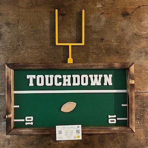 May include: A framed football-themed artwork with a green field, white yard lines, and the word "TOUCHDOWN" in bold white letters. A wooden football and a yellow goalpost are also featured. The frame is made of dark wood.