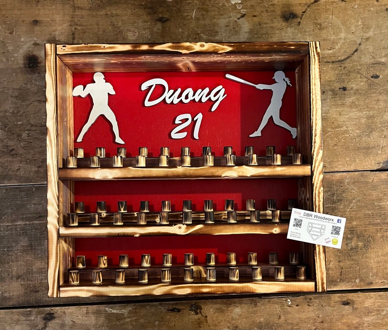 May include: A wooden baseball cap display with three shelves. The shelves are filled with baseball caps. The display has a red background with the name "Duong 21" written in white. There are two white silhouettes of baseball players on the display.