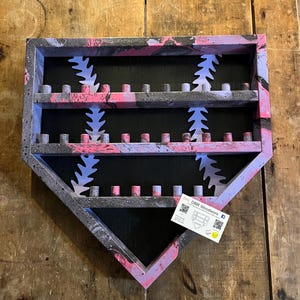 May include: A baseball-shaped wooden display shelf with a black interior and a pink, gray, and white splatter paint design. The shelf has three rows of small, cylindrical pegs for holding items. White baseball stitching is visible.