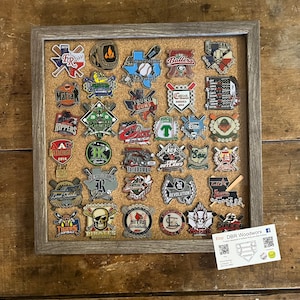 May include: A corkboard with a grey frame displays a collection of baseball-themed pins. The pins feature various team names, logos, and mascots. A business card for DBR Woodworx is displayed below the corkboard.