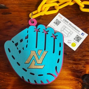 May include: A teal blue baseball glove with a gold "N" logo. The glove is attached to a yellow chain and a pink key ring. The image also includes a business card for DBR Woodworx, which specializes in custom baseball and softball ring and ball holders.