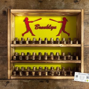 May include: A wooden baseball display shelf with two shelves and a yellow background. The shelves are filled with wooden baseball caps. The background has two red silhouettes of baseball players with bats and the name "Brooklyn" written in red letters.