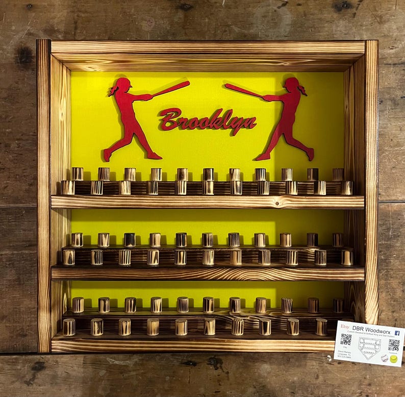 May include: A wooden baseball display shelf with two shelves and a yellow background. The shelves are filled with wooden baseball caps. The background has two red silhouettes of baseball players with bats and the name "Brooklyn" written in red letters.