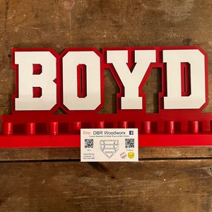 May include: A red and white wooden baseball display with the name "BOYD" in large block letters. The display has slots for baseballs and a business card with contact information.