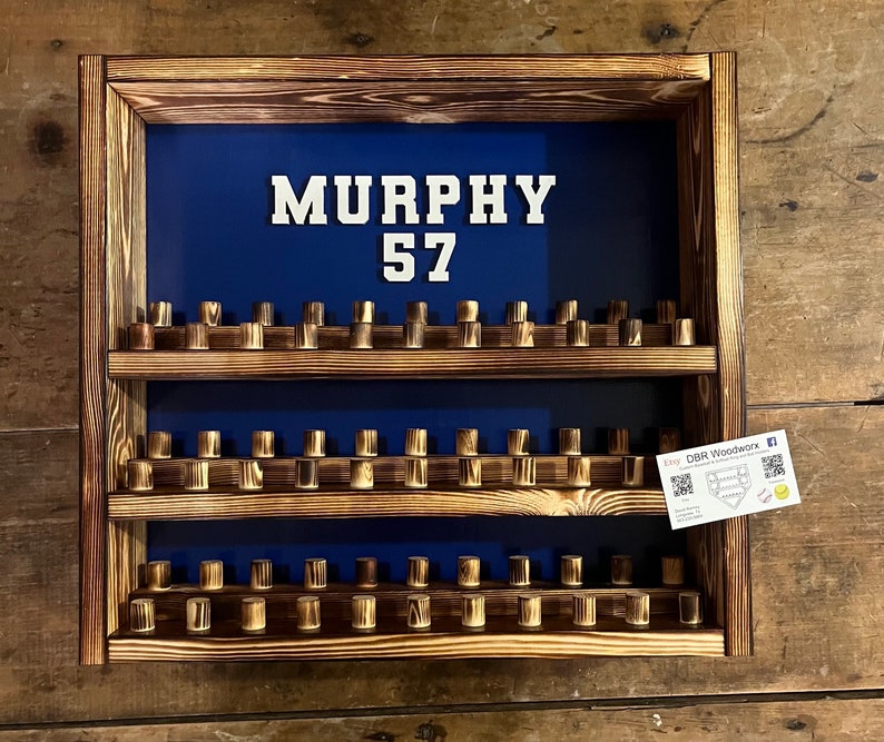 May include: A wooden baseball bat display shelf with three tiers. The shelf is painted blue and has a white sign with the name "MURPHY" and the number "57".