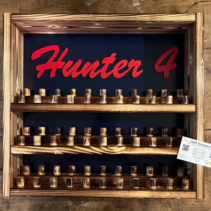 May include: Wooden wall-mounted display shelf with three tiers, featuring a dark blue background with the text "Hunter 4" in red lettering. The shelf is filled with small wooden dowels.
