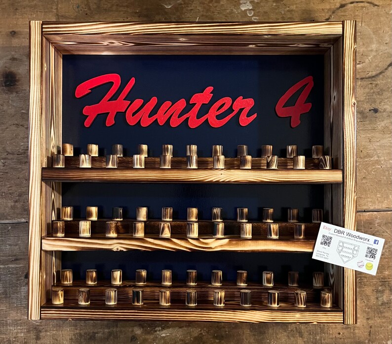 May include: Wooden wall-mounted display shelf with three tiers, featuring a dark blue background with the text "Hunter 4" in red lettering. The shelf is filled with small wooden dowels.