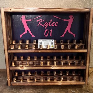 May include: A wooden baseball bat display with three shelves. The top shelf has a black background with two pink silhouettes of baseball players swinging bats. The text "Kylee 01" is written in pink on the background. The bottom two shelves have rows of wooden dowels for holding baseballs.