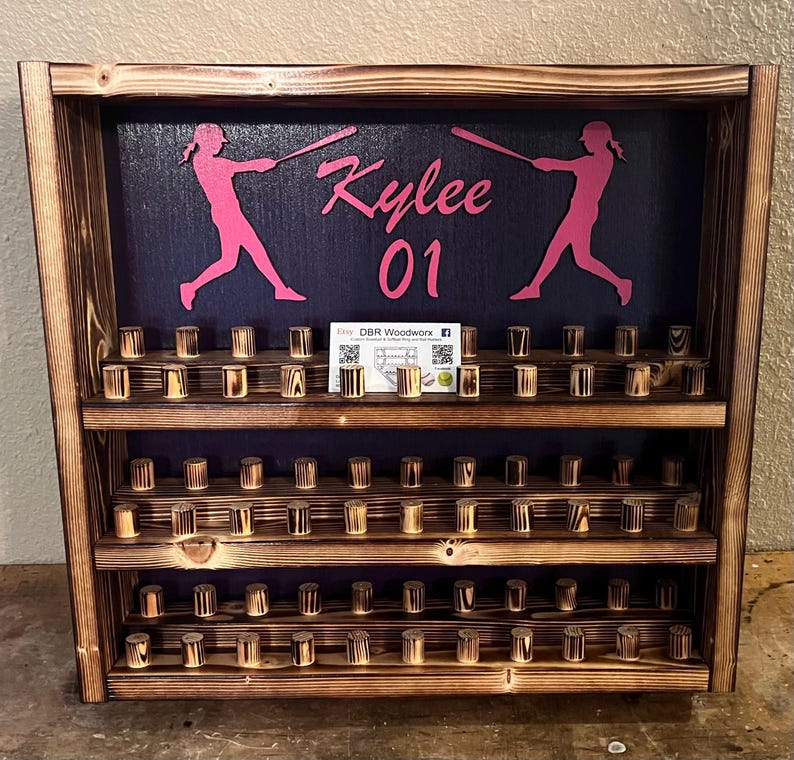 May include: A wooden baseball bat display with three shelves. The top shelf has a black background with two pink silhouettes of baseball players swinging bats. The text "Kylee 01" is written in pink on the background. The bottom two shelves have rows of wooden dowels for holding baseballs.