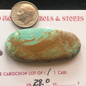 May include: An oval turquoise and brown cabochon gemstone with a dime for size comparison. The gemstone has a polished surface and a unique pattern. The dime is dated 1994. The text "BOLS & STONES" is visible.