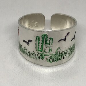May include: Silver adjustable ring with a green cactus, green grass, and black birds on a white background.