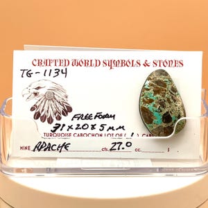 May include: A turquoise cabochon with brown matrix, displayed on a card with the text "Crafted World Symbols & Stones" and "Apache". The stone measures 31x20x5 mm. The card also features an eagle illustration.