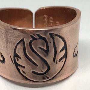 May include: A copper-colored adjustable ring with a central black design featuring the letter "S" within a stylized leaf shape. The ring has etched details and a brushed finish, with additional small designs.