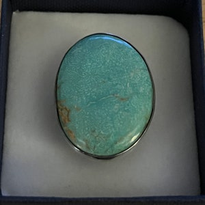 May include: Oval turquoise gemstone ring with a silver-colored metal band, presented in a dark blue gift box with a light gray interior. The turquoise stone has a mottled pattern and a polished surface.