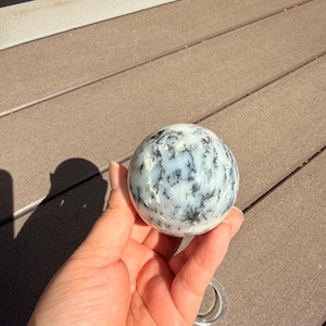 May include: A spherical, decorative stone with a white and light blue base color, and black dendritic patterns. The stone is held in a hand, and the background is a brown wooden deck.