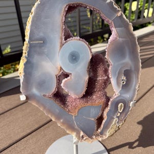 May include: A large, oval-shaped agate slice featuring translucent light gray and blue banding surrounding a central cluster of deep purple amethyst crystals. The mineral specimen is mounted on a circular white metal stand for display.