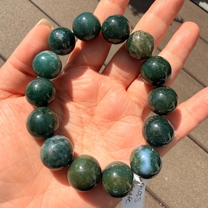 May include: A bracelet made of large, round moss agate beads. The beads are primarily dark green with lighter green and white inclusions. The bracelet is held in a hand, showcasing the polished, natural stone. The beads appear to be approximately 1 inch in diameter.