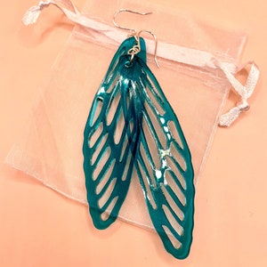 May include: Teal-colored, wing-shaped earrings with intricate cut-out designs. The earrings are suspended from silver-colored hooks and rest on a sheer, light pink pouch. The background is a solid, light peach color.