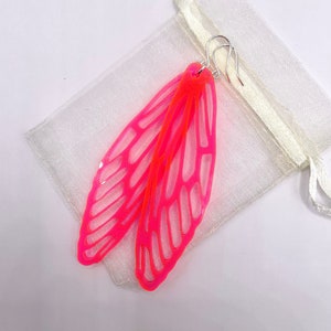 May include: A pair of neon pink and orange wing-shaped earrings with silver hooks. The earrings are made of a translucent material and are displayed on a white background next to a small white pouch.