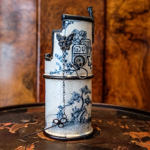 Puede incluir: Una pipa de opio cilíndrica de cerámica vintage con un diseño floral y paisajístico blanco y azul. La pipa presenta un borde de metal negro, un adorno en forma de mariposa y una cadena. Una herramienta de metal descansa junto a la pipa.