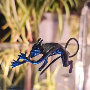 May include: A dark blue figurine of a creature with horns, a long tail, and outstretched arms. The figurine is posed in a dynamic, almost leaping position. The background is blurred, with green plants visible.